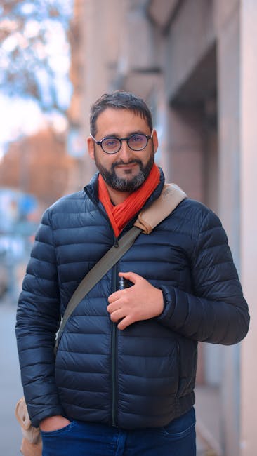 Outdoor portrait of a smiling man with glasses in warm clothing, standing on a city sidewalk.